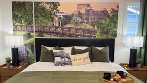 Et hotelværelse med en seng, et natbord med en lampe, en bakke med en doughnut og kaffekopper, samt en væg med et bybillede-plakat.