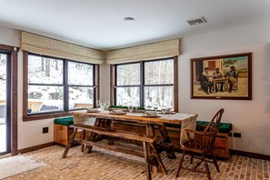 Un coin repas avec une table en bois dressée pour quatre personnes, de grandes fenêtres avec des rideaux et un tableau accroché au mur.
