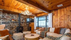 A cozy cabin interior with a stone wall, a fireplace, a television, and a large window. - Lux Hot Tub Cabin w Fire Pit (Napanoch)