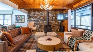 A cozy living room with a stone fireplace, leather sofa, and a large window. - Lux Hot Tub Cabin w Fire Pit (Napanoch)