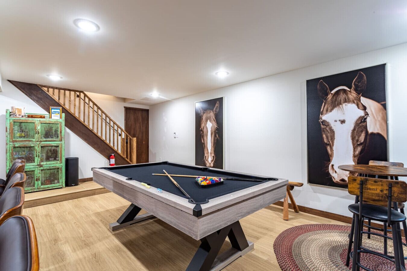 A pool table in a room with a staircase, a green cabinet, and two horse-themed paintings.
