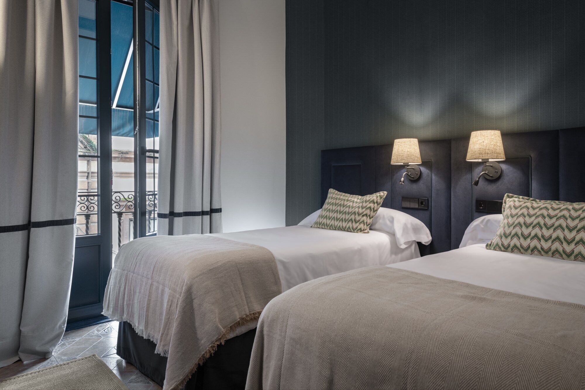 A hotel room with two beds, a large window with curtains, and wall-mounted lamps.