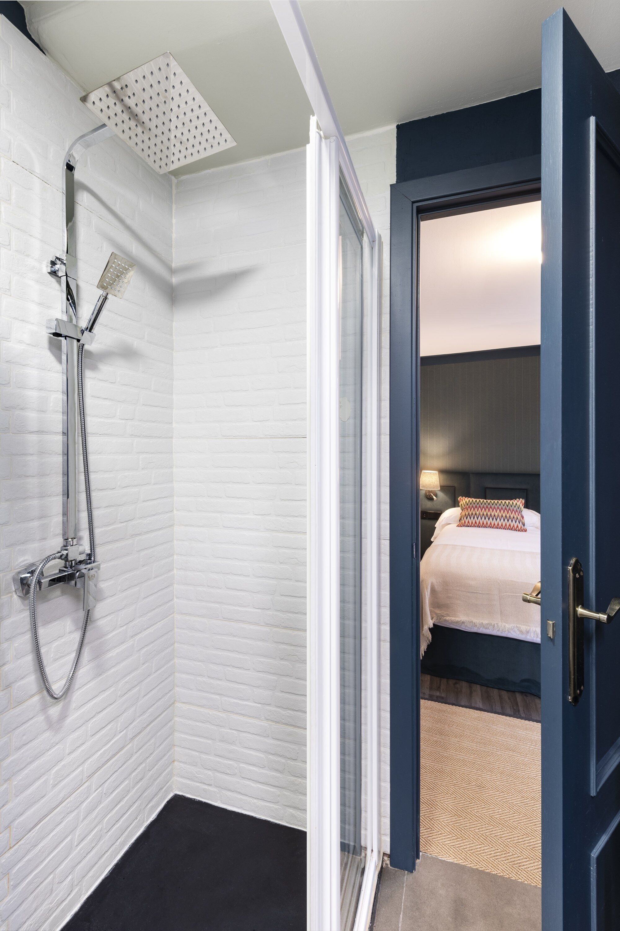 Un baño en esquina con ducha, pared de ladrillo blanco y puerta que da a un dormitorio con cama y mesita de noche.