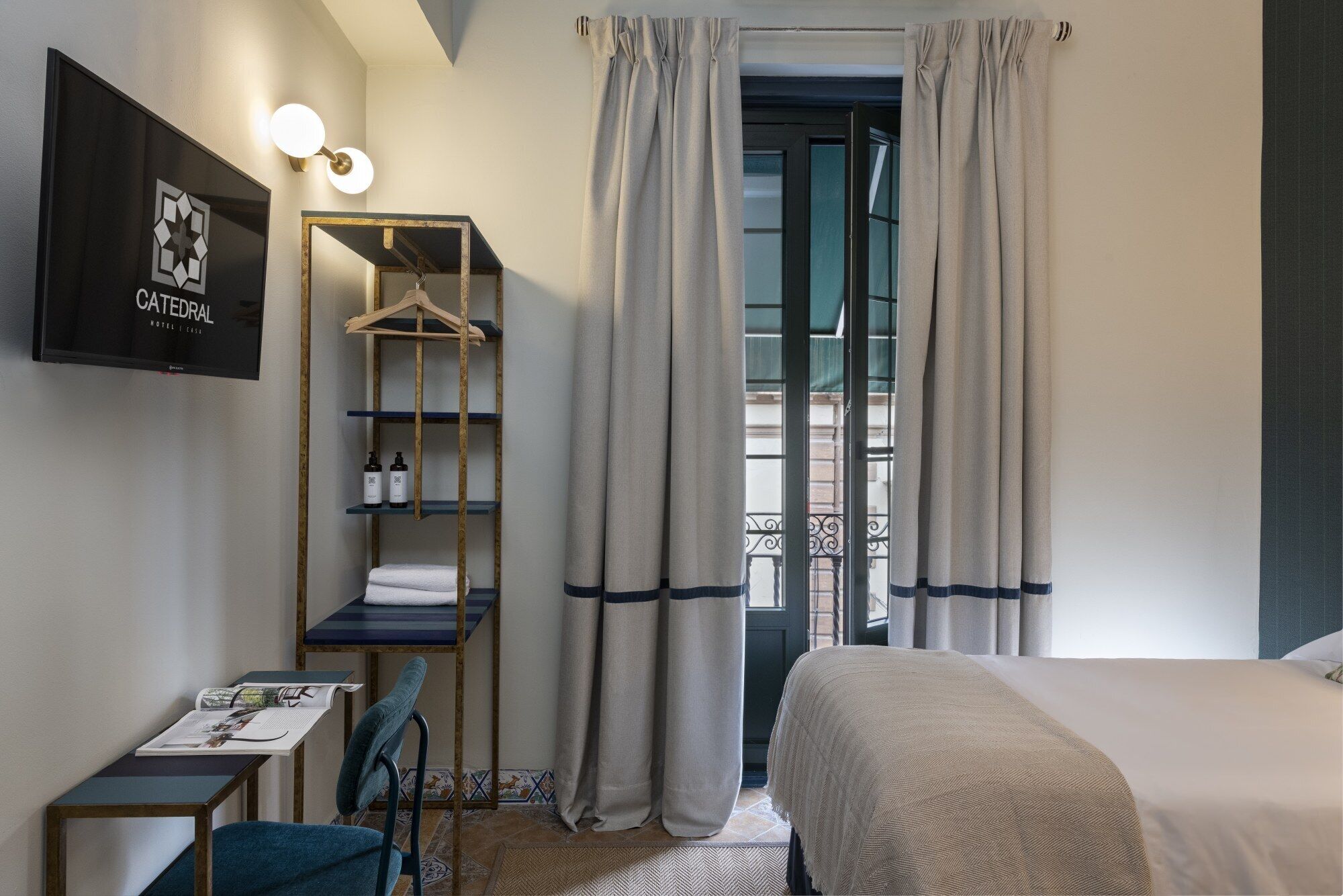 A hotel room with a bed, a desk with a chair, a hanging shelf with toiletries, and a view through a window with curtains.