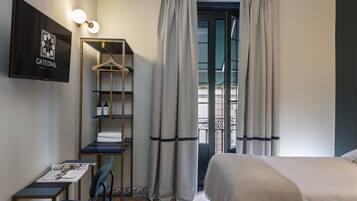 A hotel room with a bed, a desk with a chair, a hanging shelf with toiletries, and a view through a window with curtains.