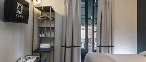 A hotel room with a bed, a desk with a chair, a hanging shelf with toiletries, and a view through a window with curtains.