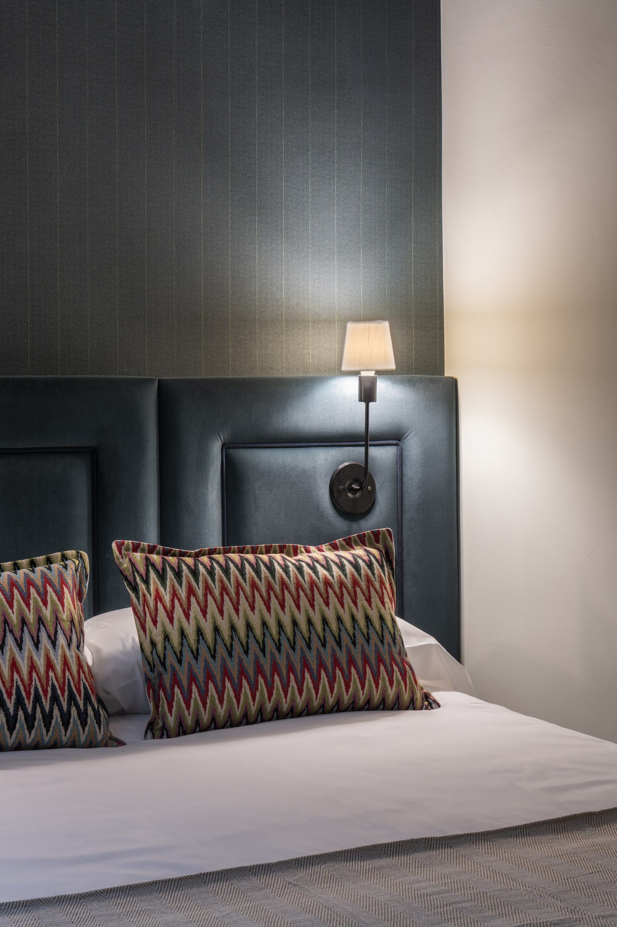 Una cama con cabecera, dos almohadas estampadas y una lámpara fijada a la pared.