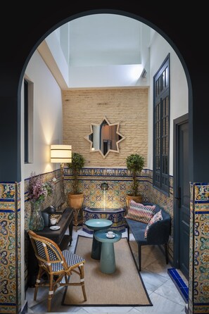 An interior courtyard with a tiled wall, a wooden bench, a table with chairs, and a potted plant.