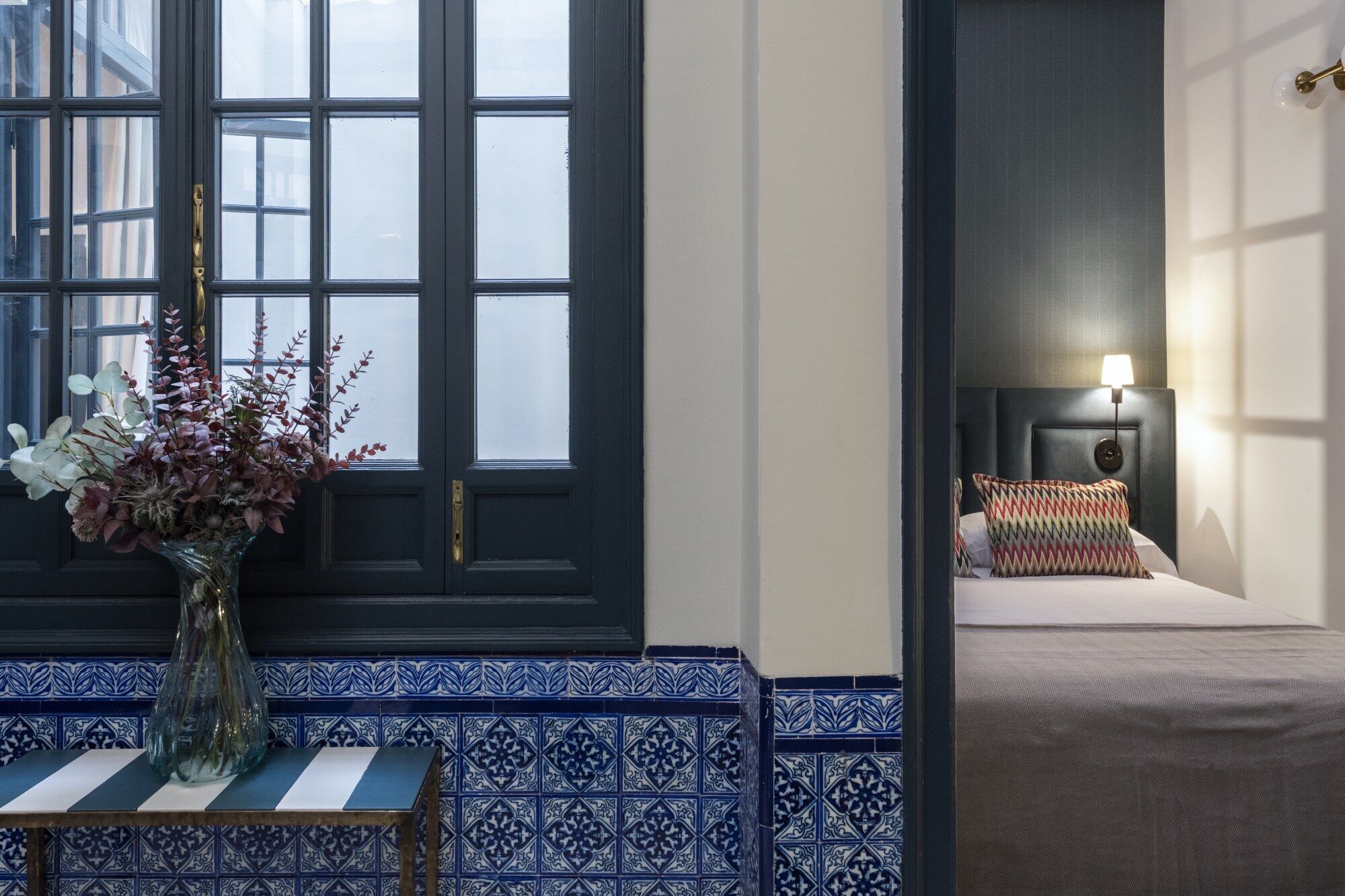 A hotel room with a large window, a bedside table with a vase of flowers, a bed with a patterned pillow, and a headboard with a wall-mounted lamp.