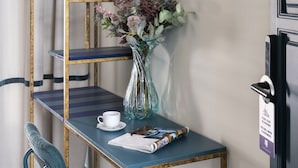 Une pièce avec une chaise bleue, une petite table sur laquelle est posé un vase de fleurs, une bibliothèque et un luminaire fixé au mur.