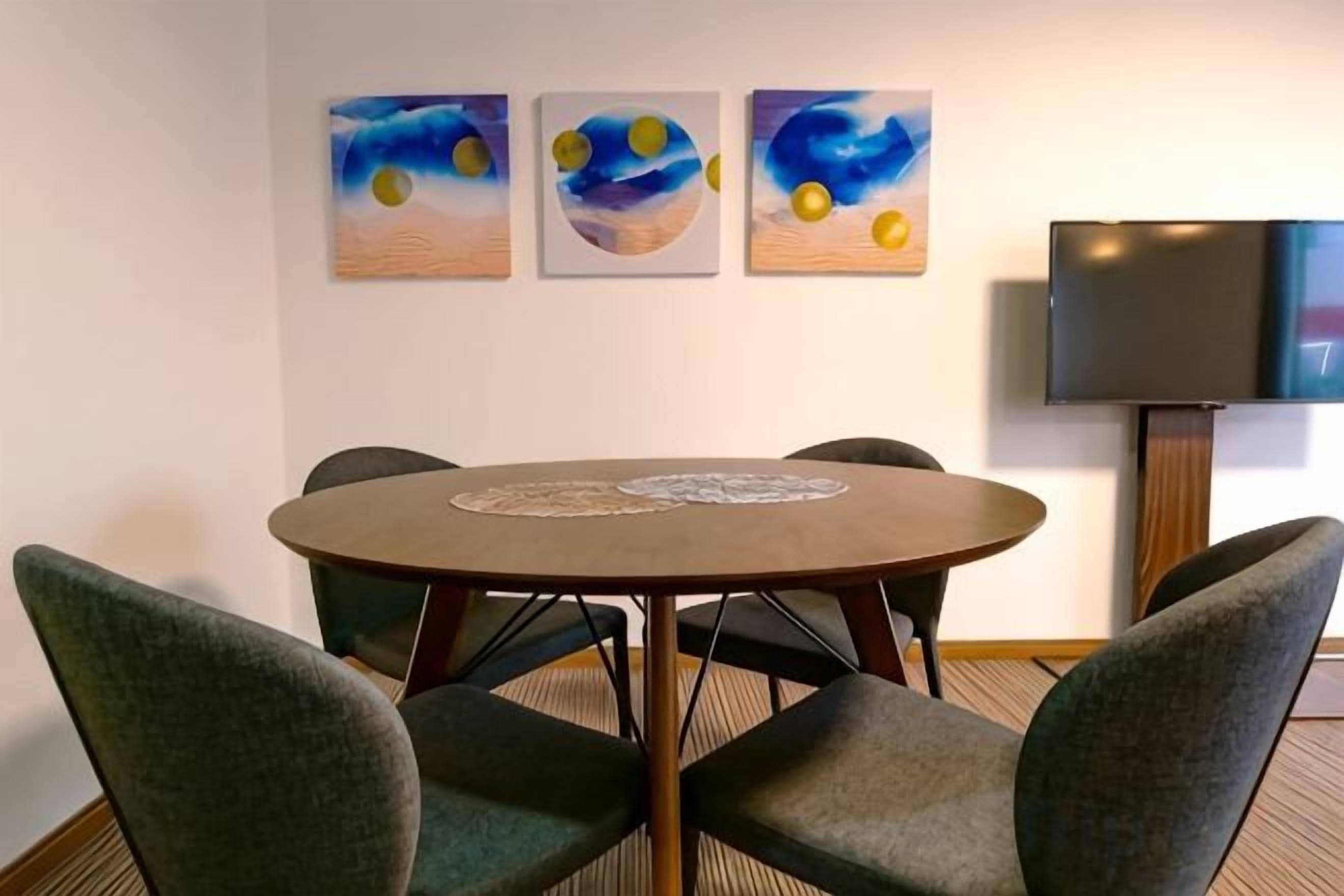 A conference room with a round table, four chairs, and three abstract paintings on the wall.