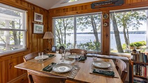 A wooden dining table set with plates, glasses, and cutlery, with a view of a lake through large windows.