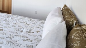 A bedroom with a bed, white floral bedding, a wall-mounted air conditioner, and a bedside lamp.