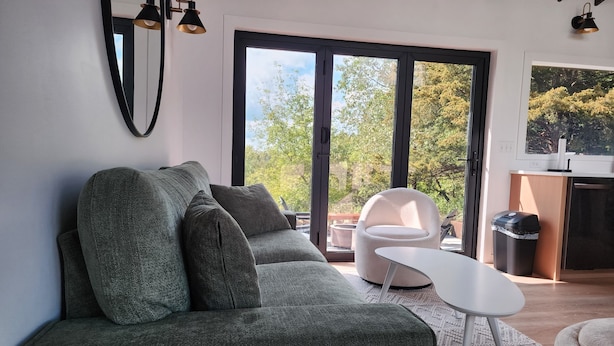 A modern living room with a grey sofa, a white side table, and a white chair. There is a large window with a view of trees and a kitchen area with a trash bin and a sink.