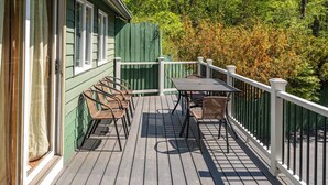 Une terrasse en bois avec une table et des chaises, entourée d’une balustrade blanche, et une clôture verte en arrière-plan.