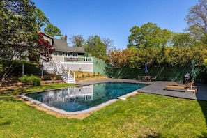 A backyard with a swimming pool, a wooden deck, and a fenced area with a house in the background.