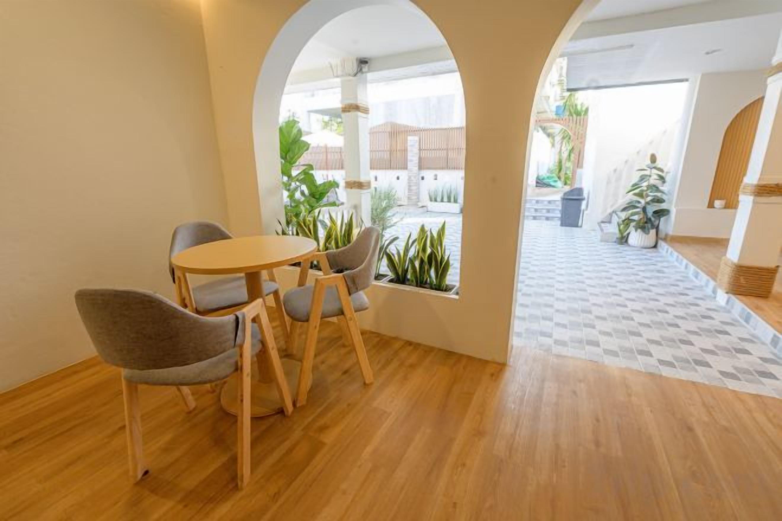 A dining area with a round table and four chairs, a wooden floor, and a view through an archway to an outdoor space with plants and a building.