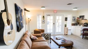 A cozy living area with a sofa, a coffee table, and a guitar. There is a door with an exit sign, a small table with a plant, and a kitchenette in the background. - Desert Dream Airstream with Pool (Yucca Valley)