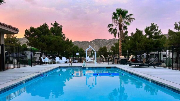 A rectangular swimming pool with a clear blue water surface, surrounded by a paved deck. There are lounge chairs and a small gazebo-like structure in the background. - Desert Dream Airstream with Pool (Yucca Valley)