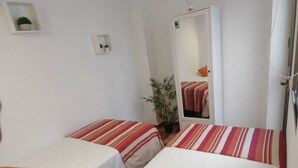 Une chambre d’hôtel avec deux lits simples, chacun recouvert d’une couvre-lit à rayures et agrémenté d’un oreiller orange. Une plante en pot est placée entre les lits, et un miroir sur le mur reflète l’image de la pièce.