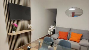 Un salon moderne avec une table à manger en verre, deux chaises bleues, un canapé gris avec des coussins orange, un téléviseur à écran plat, un miroir rond et une orchidée en pot.