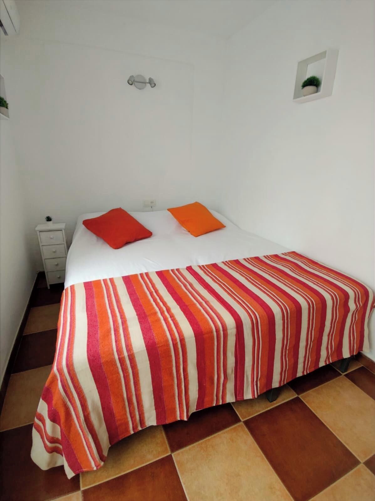 Une chambre avec un lit simple, une couvre-lit à rayures, deux oreillers, une petite table de chevet et un luminaire fixé au mur.