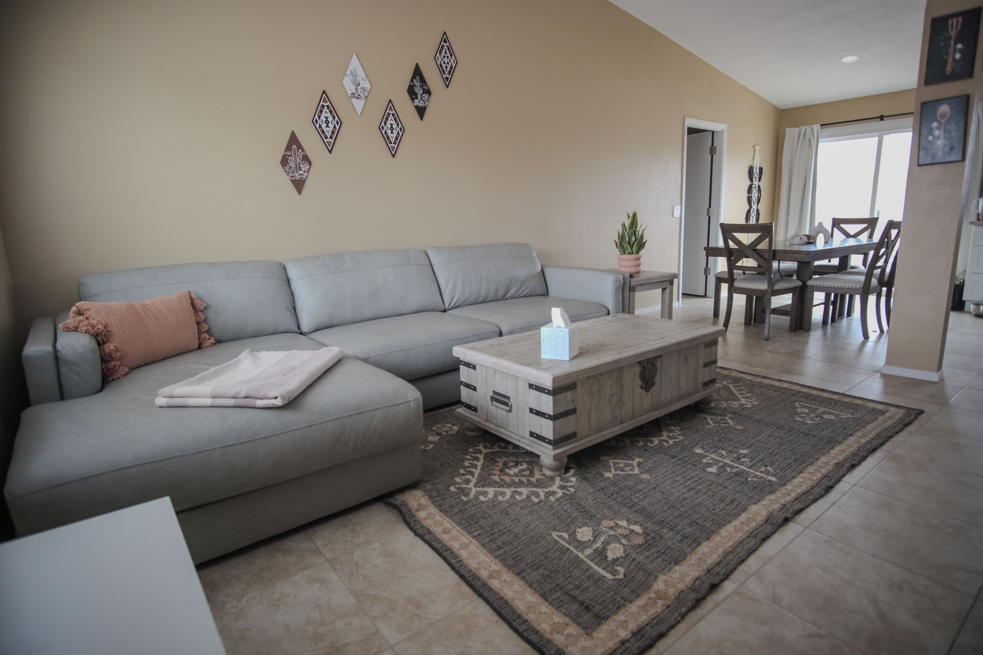 A living room with a grey sofa, a wooden coffee table, and a patterned rug. There is a dining area with a table and chairs visible in the background.