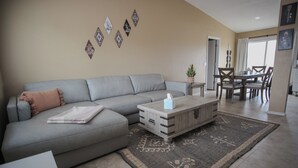 Una sala de estar con un sofá gris, una mesa de centro de madera y un tapete estampado. Se ve un comedor con una mesa y sillas al fondo.