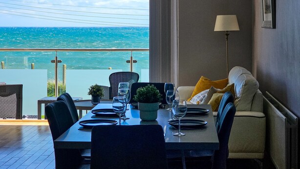 Un coin repas avec une table dressée pour quatre personnes, un canapé avec des coussins, une lampe sur pied et une vue sur la mer.