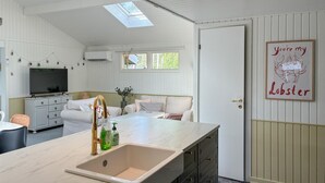 Ett väl upplyst rum med ett stort fönster, en vit soffa, ett litet bord och en diskho med en bänkskiva i marmor.