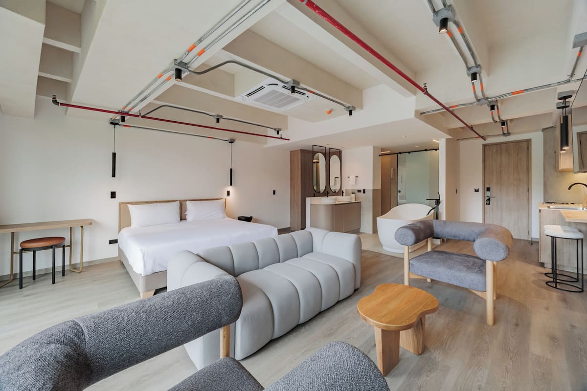 A modern hotel room with a large bed, grey sofas, a wooden table, and a bathroom with a mirror and sink.