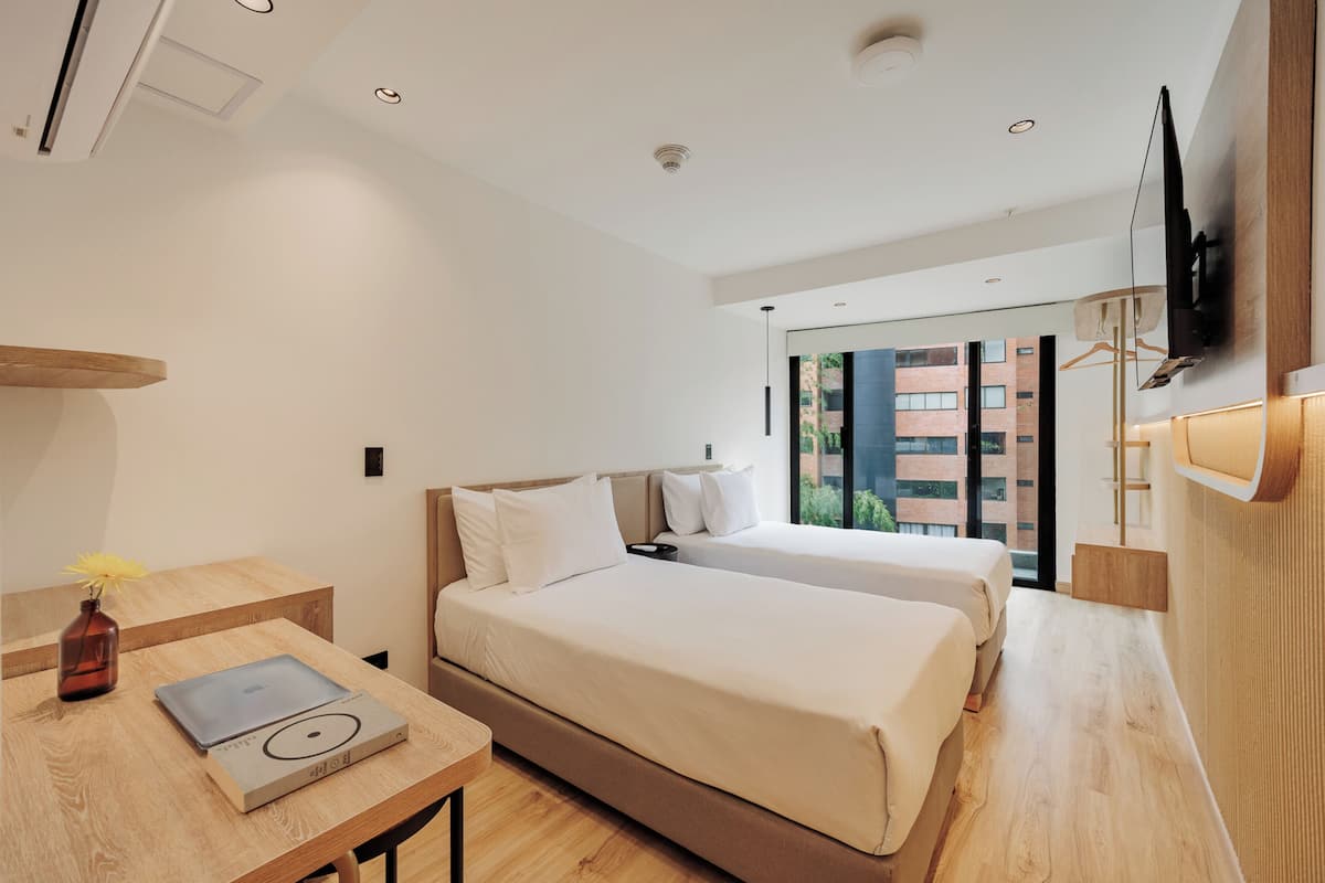 A modern hotel room with two beds, a desk with a laptop, a vase with a flower, and a TV mounted on the wall.