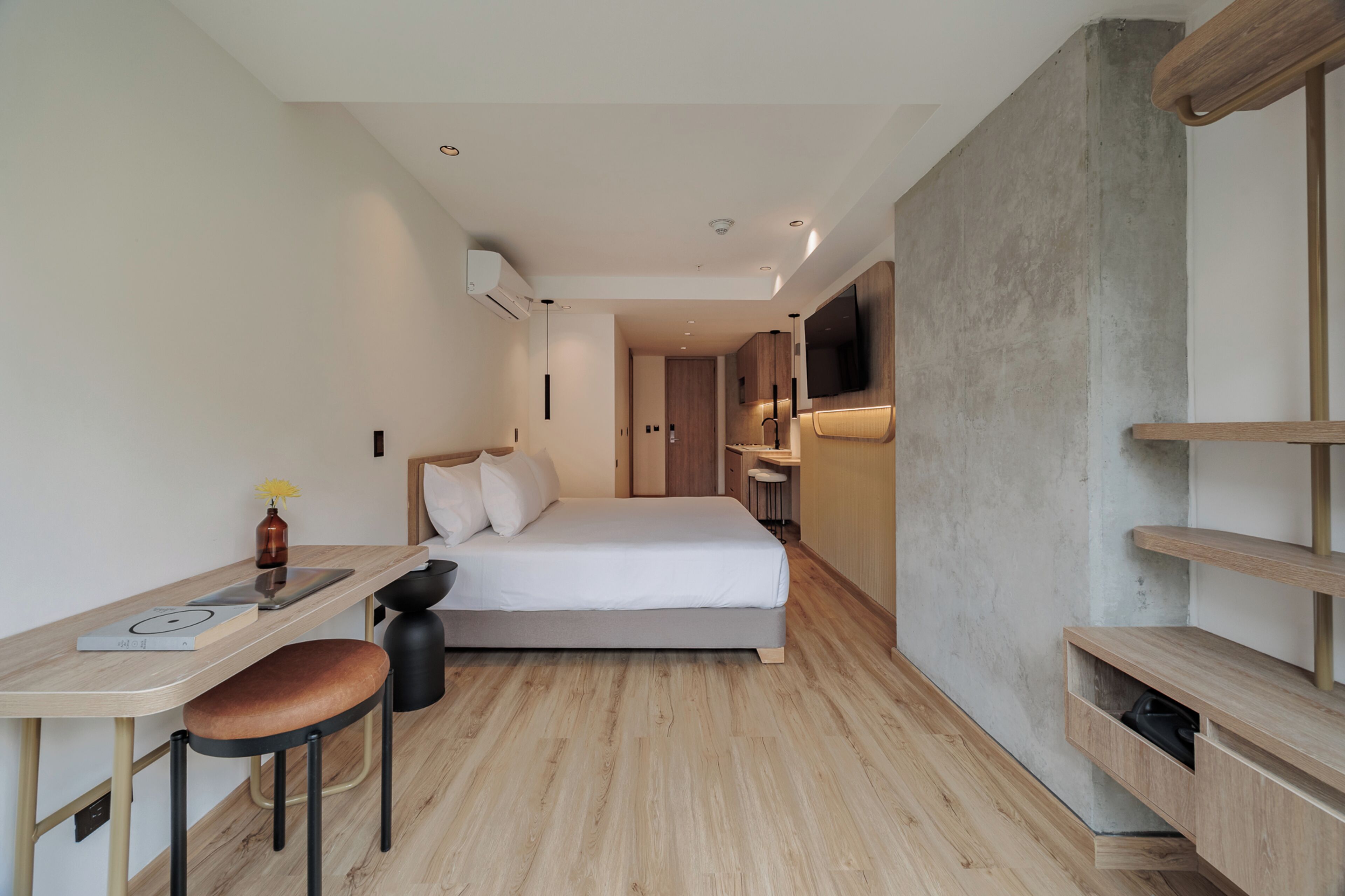 A modern hotel room with a bed, a desk with a chair, a TV mounted on the wall, and a small seating area with stools.