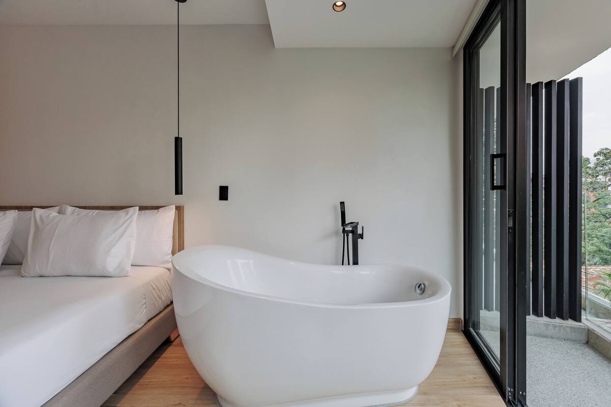 A modern bedroom with a large freestanding bathtub, a bed with white linens, and a view through a glass door.