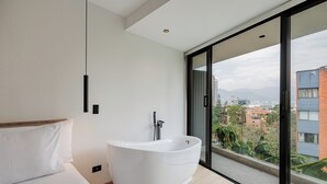 A modern bathroom with a freestanding bathtub, a bed with white linens, and a large window offering a city view. - Lomm By Somos The Group (Medellín)