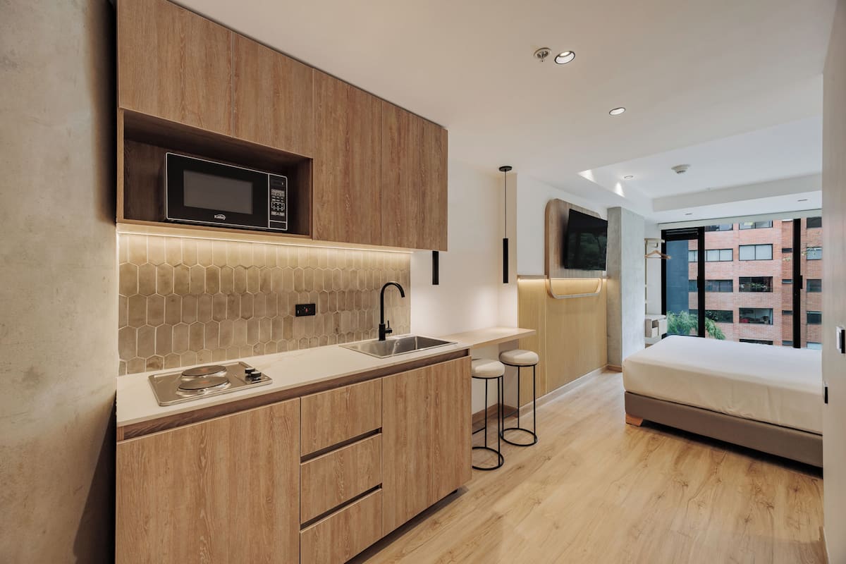 A modern kitchen with wooden cabinets, a microwave, and a tiled backsplash. There are stools and a bed in the adjacent room.