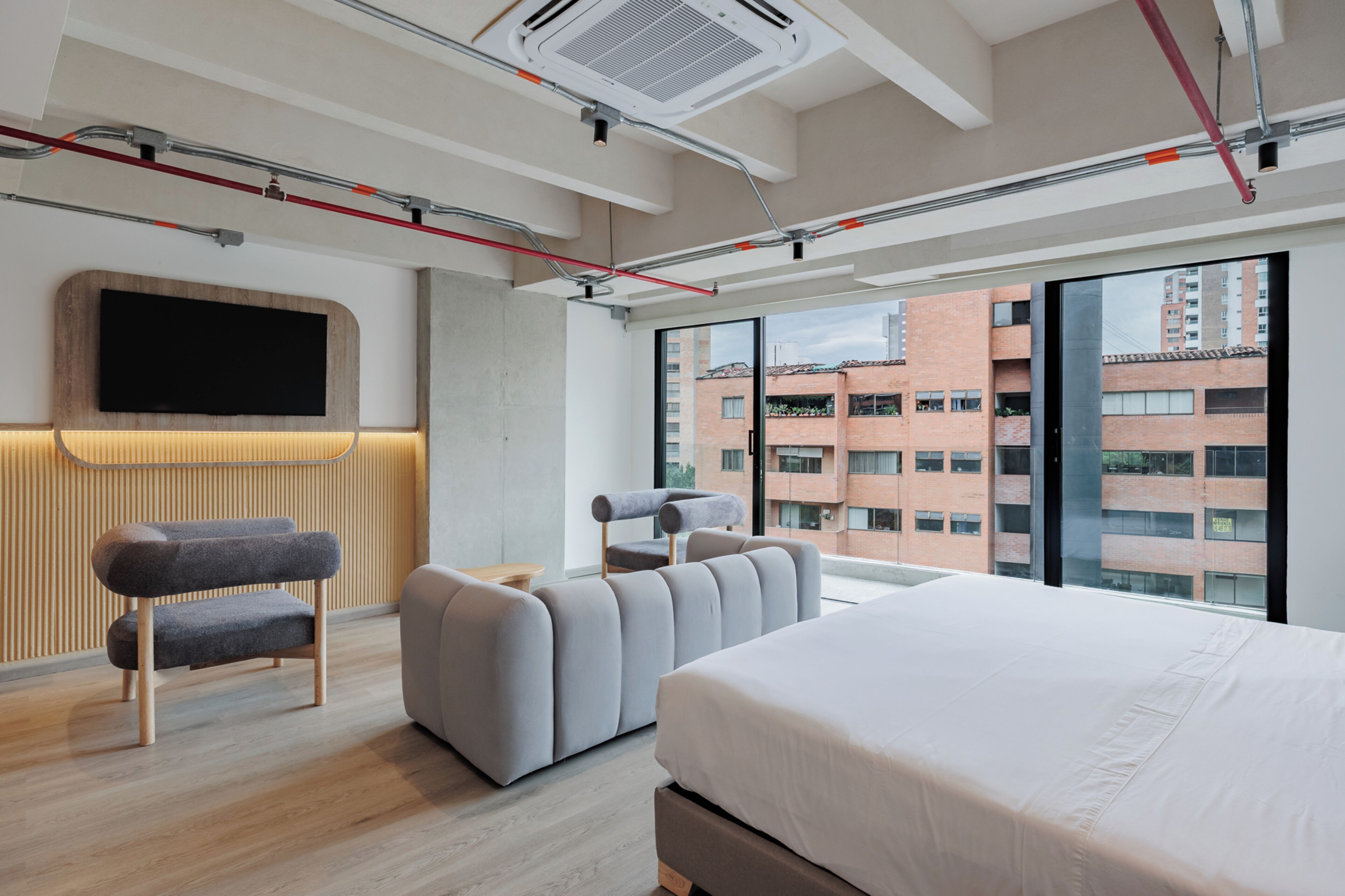 A modern hotel room with a large bed, a sofa, a TV mounted on the wall, and a view of the cityscape through large windows.