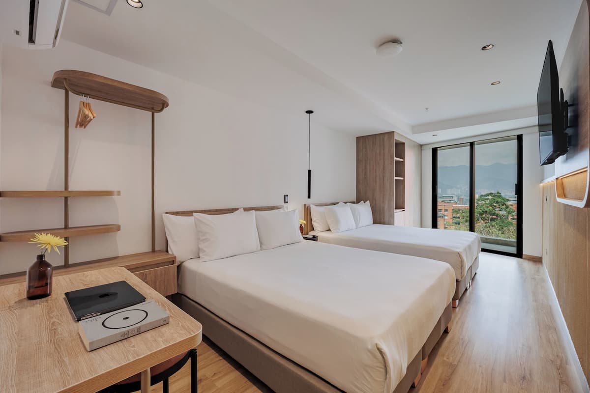 A modern hotel room with two beds, a wooden desk with a laptop, a vase with flowers, and a large window offering a city view.