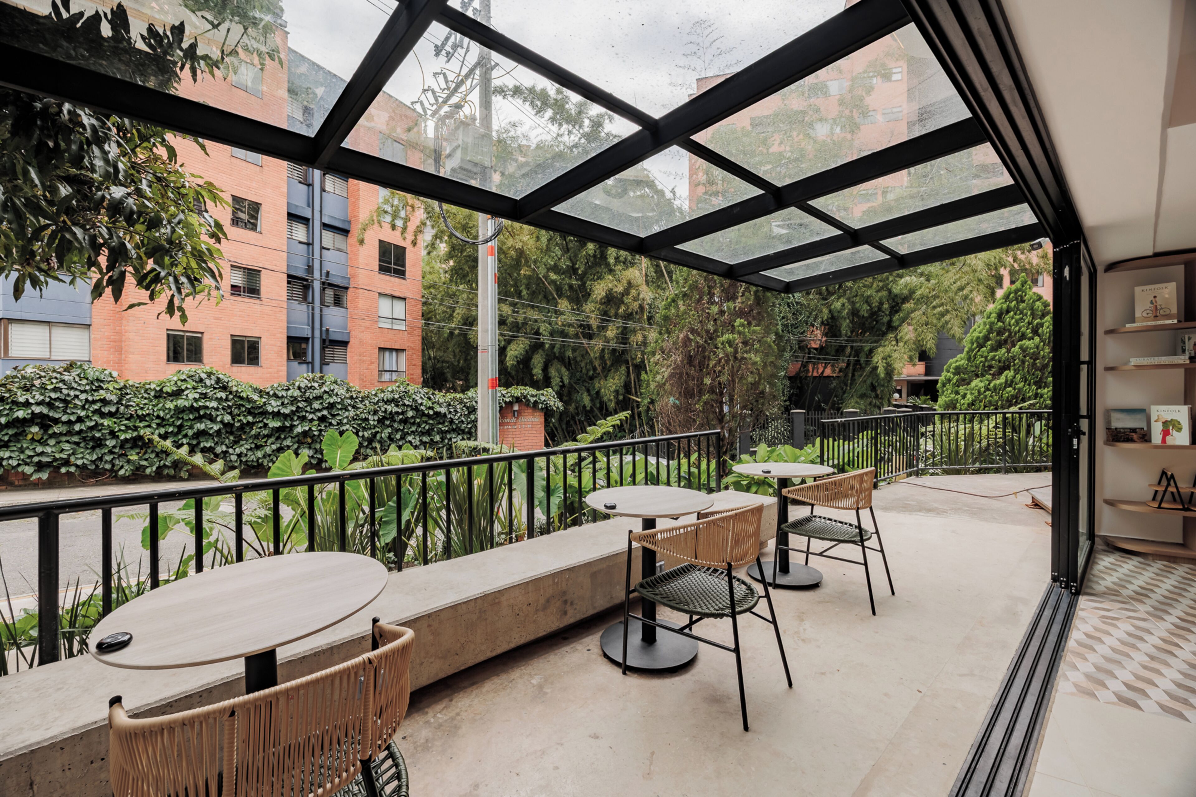An outdoor patio with tables and chairs, a glass ceiling, and a view of buildings and greenery.