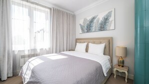 A bedroom with a large bed, a bedside table with a lamp, a window with sheer curtains, and a wall-mounted artwork. - Toompea Castle 2 BR Old Town Apartment (Tallinn)
