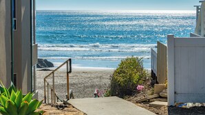 A coastal property with a view of the ocean, a concrete pathway leading to the beach, and a white fence. - Beach Bum Retreat (Cayucos)