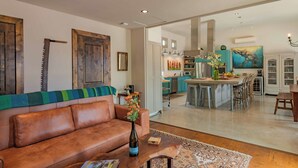 A living room with a brown leather sofa, a coffee table with a vase, and a patterned rug. A kitchen with a central island and bar stools is visible through an open doorway. - Refugio Cosetta (Paso Robles)
