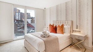 A modern bedroom with a large bed, a view of buildings through floor-to-ceiling windows, and a minimalist decor.