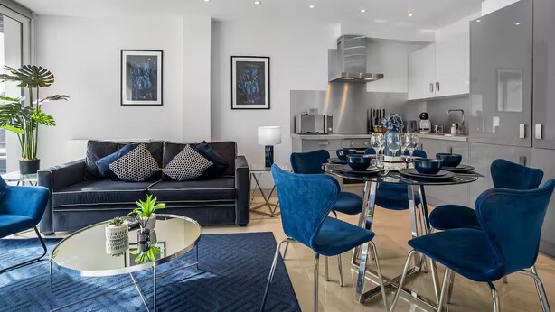 Una sala de estar moderna con un sofá, una mesa de centro y sillas azules. Hay un comedor con una mesa preparada para cuatro personas y una cocina con armarios y electrodomésticos.