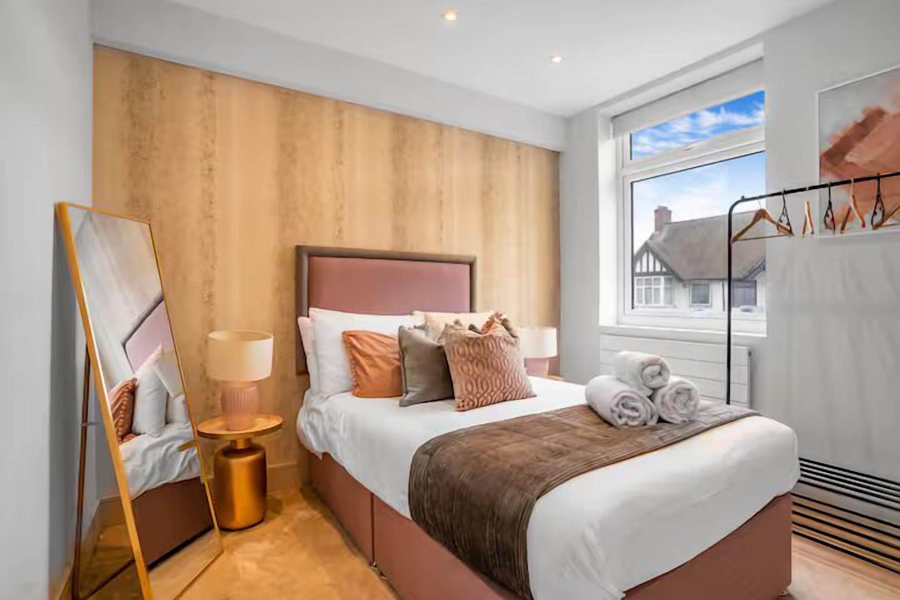 A modern bedroom with a large bed, a bedside table, a mirror, and a view of a house through a window.