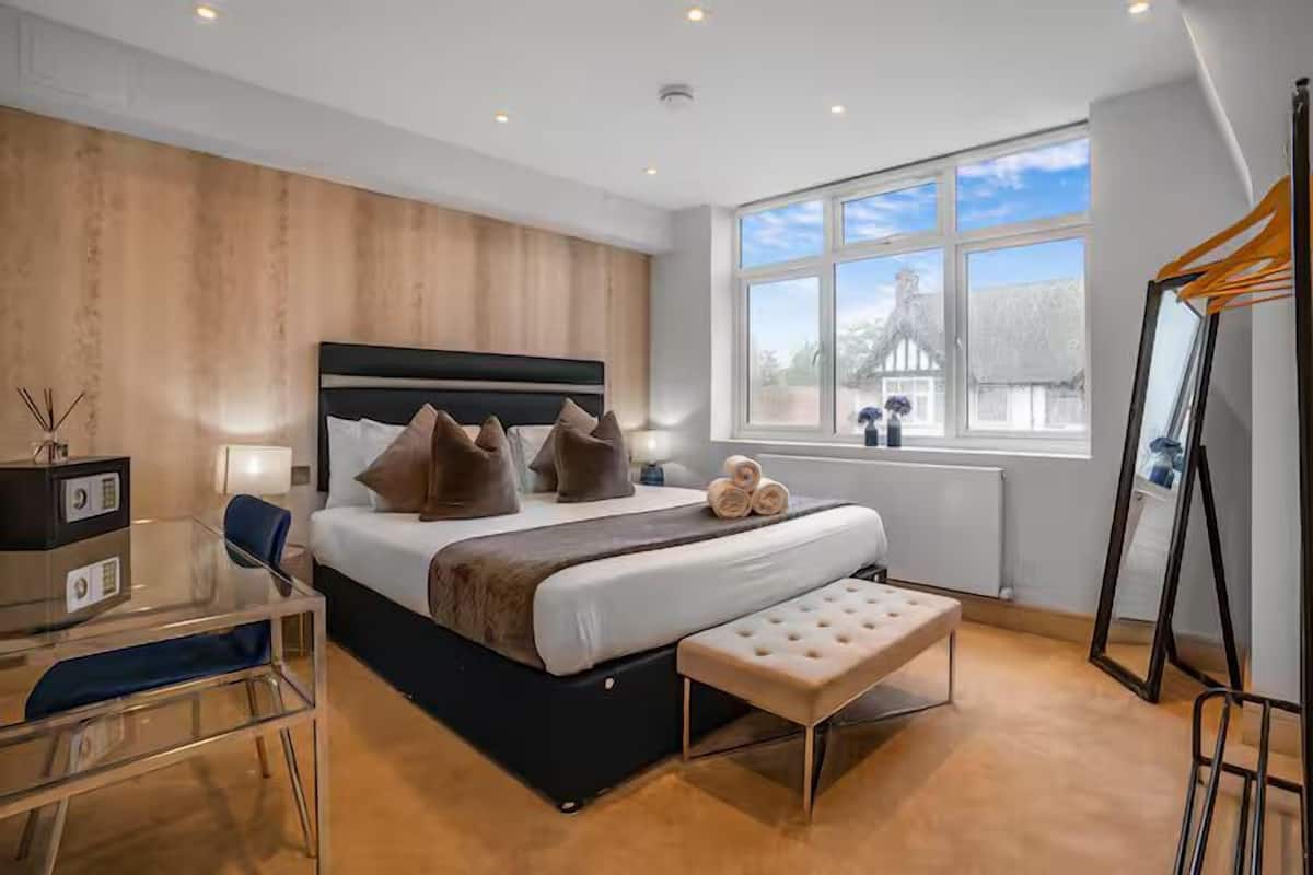 A modern hotel room with a large bed, a glass desk, a bench, and a window with a view of the outside.