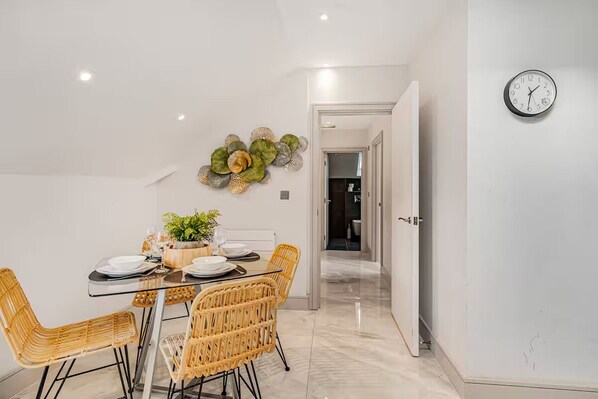 A modern dining area with a glass table, wicker chairs, and a decorative wall piece. - Wembley Park Flats | 5 mins from Station (Wembley)
