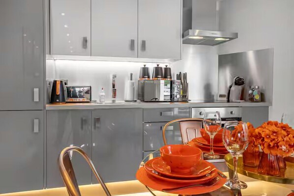 Une cuisine moderne avec des meubles gris, des appareils en acier inoxydable et un coin repas agrémenté d’assiettes orange et d’un vase de fleurs.