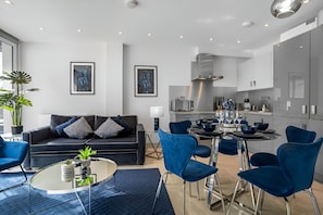 En moderne stue med sofa, sofabord og lænestole. Der er et spiseområde med et bord dækket til fire personer. Køkkenet ses i baggrunden med moderne hårde hvidevarer.