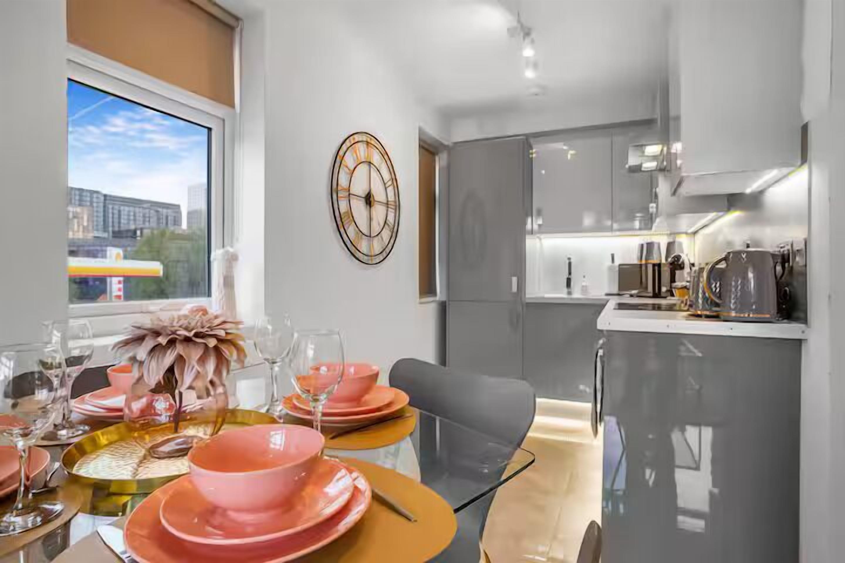 A modern kitchen with grey cabinets, a countertop with a coffee machine, and a dining area with pink plates and glasses.